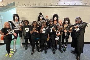Group of people wearing black tshirts standing in front on KISS, wearing glamrock makeup and outfits