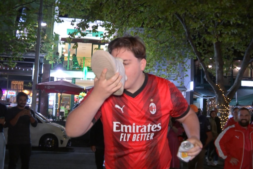 A man wears a red shirt and drinks from a white shoe on a dark Lygon Street.