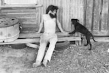 Simon sitting on a piece of wood in front of an old shed looking at his dog Merlin.