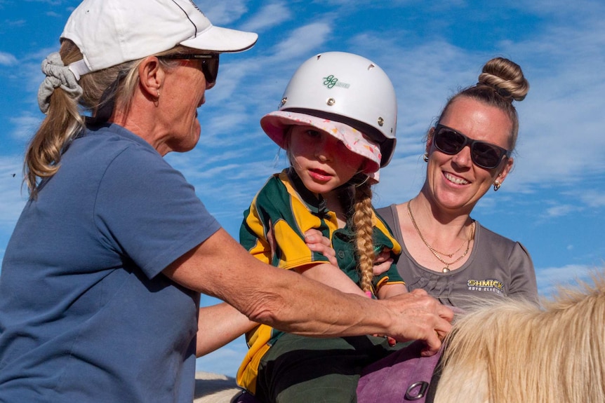 Pippa, centre, wears white helmet and school uniform. Two women help her and she is sat on a cream coloured pony.