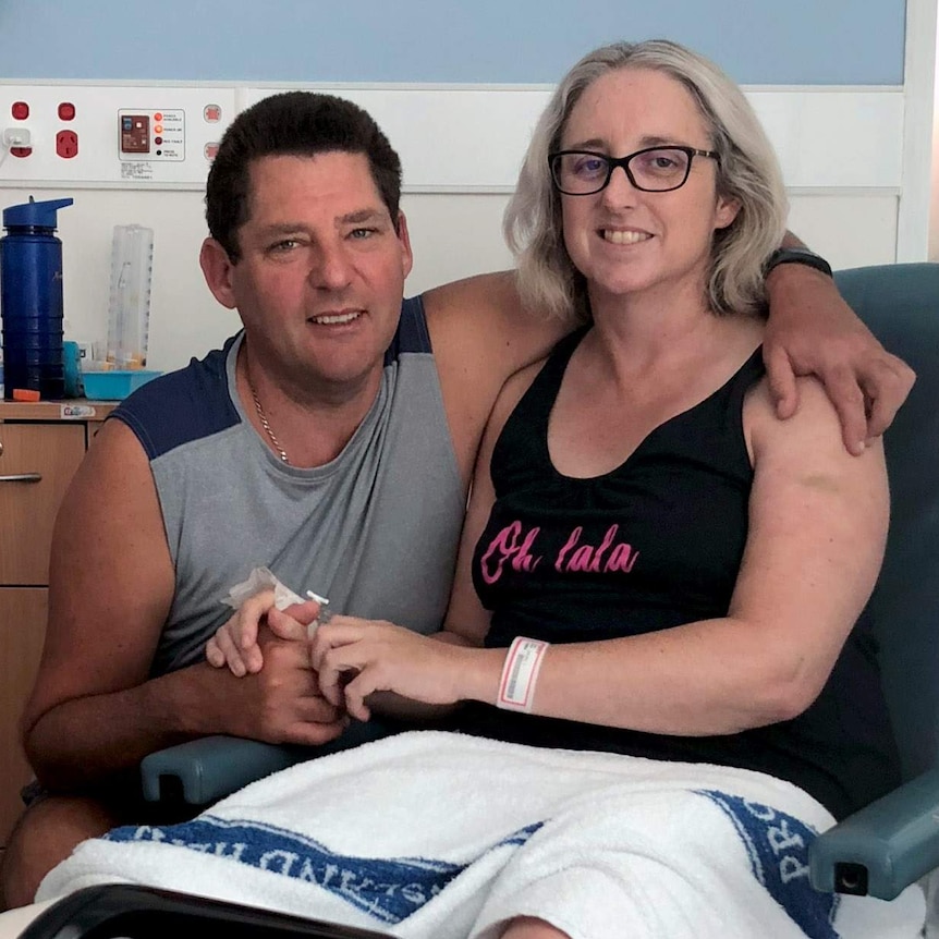 A smiling Justine Barwick sits in a hospital chair with a towel over her legs while her husband puts his arm around her.