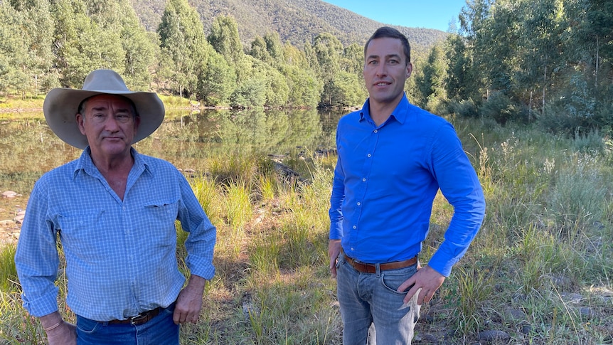 Two men in blue shirts stand with their hands on their hips along a stretch of river.