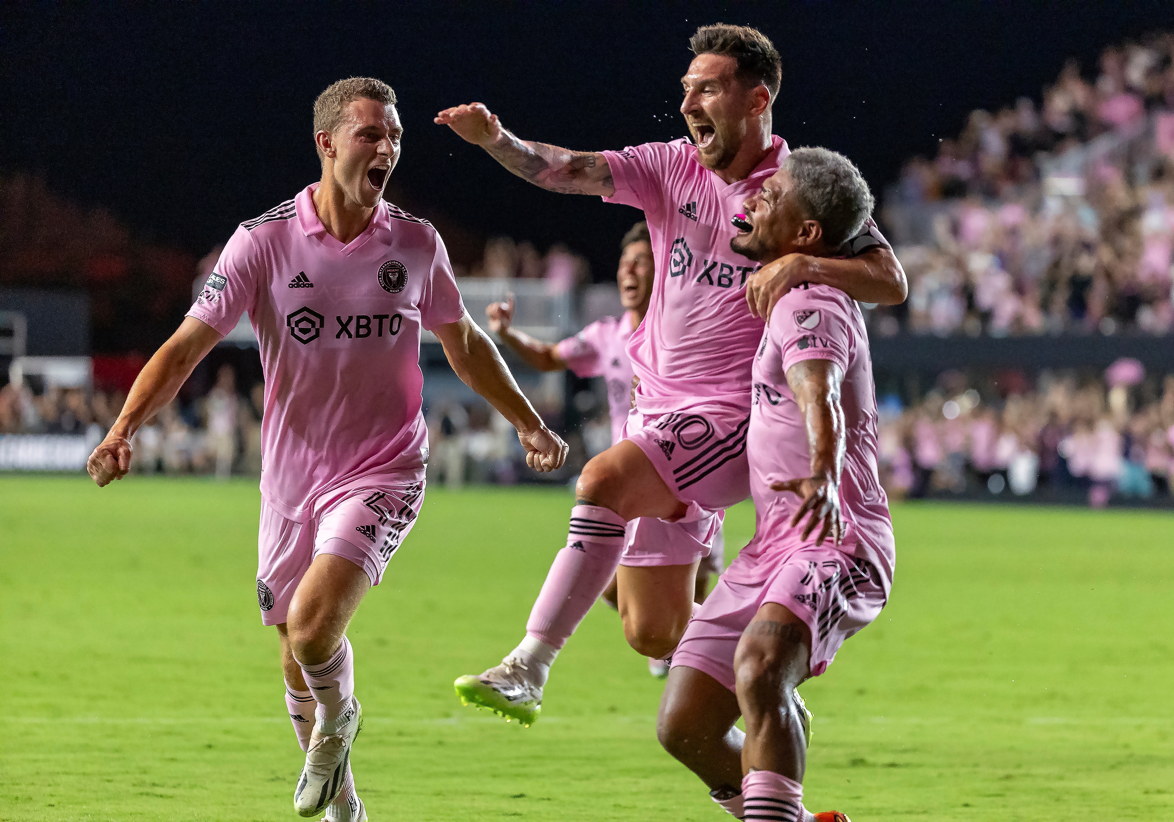 Three men in pink soccer uniforms leap into each others arms on a field 
