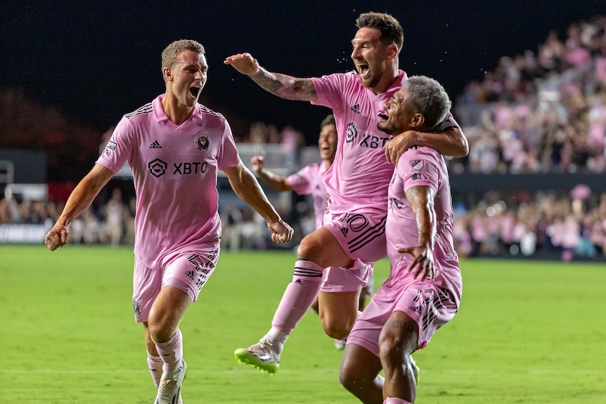 Three men in pink soccer uniforms leap into each others arms on a field 