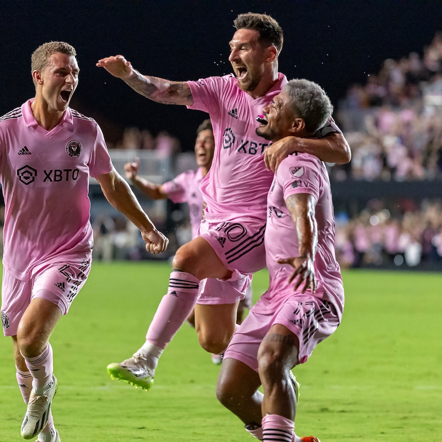 Three men in pink soccer uniforms leap into each others arms on a field 