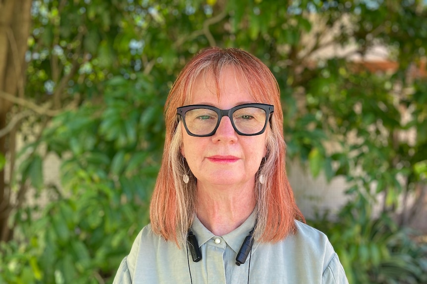 A woman wearing black glasses stands in front of some greenery.