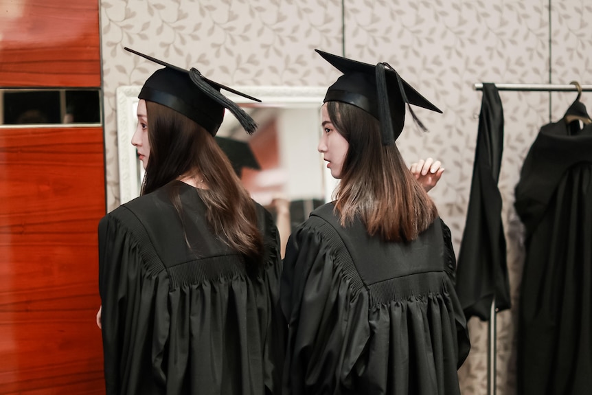 Two women in black graduation gowns and hats look over their shoulder.