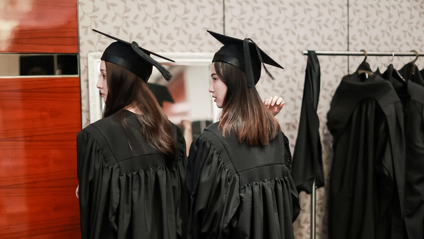 Two women in black graduation gowns and hats look over their shoulder.