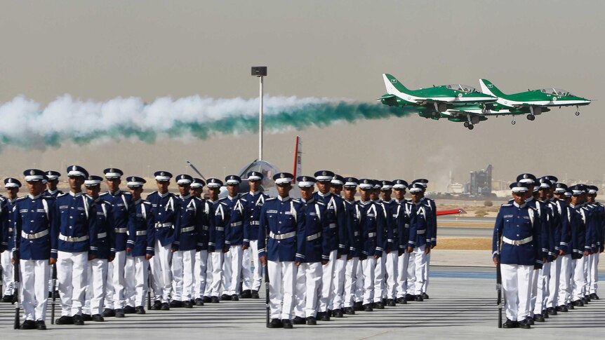 jets fly over lines of men in military uniform as green and white smoke trails behind the jets