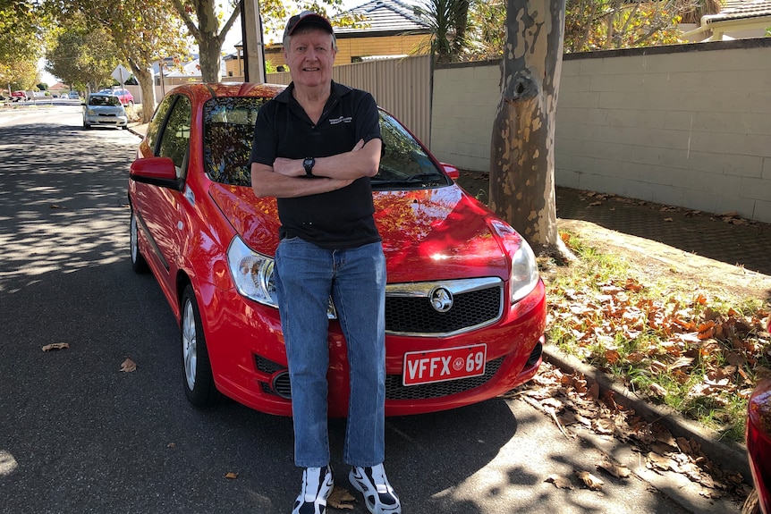 Stewart Underwood, in jeans, black shirt and cap, smiles leaning on a shiny red Holden in a leafy suburban street.