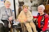 Three women gather around a 100th birthday cake with lit candles   