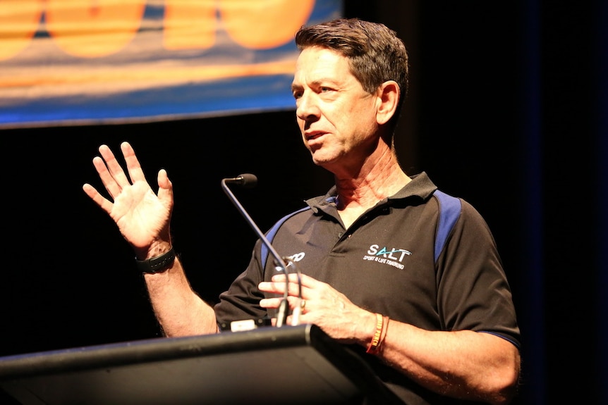 man in black top standing at microphone, speaking at event