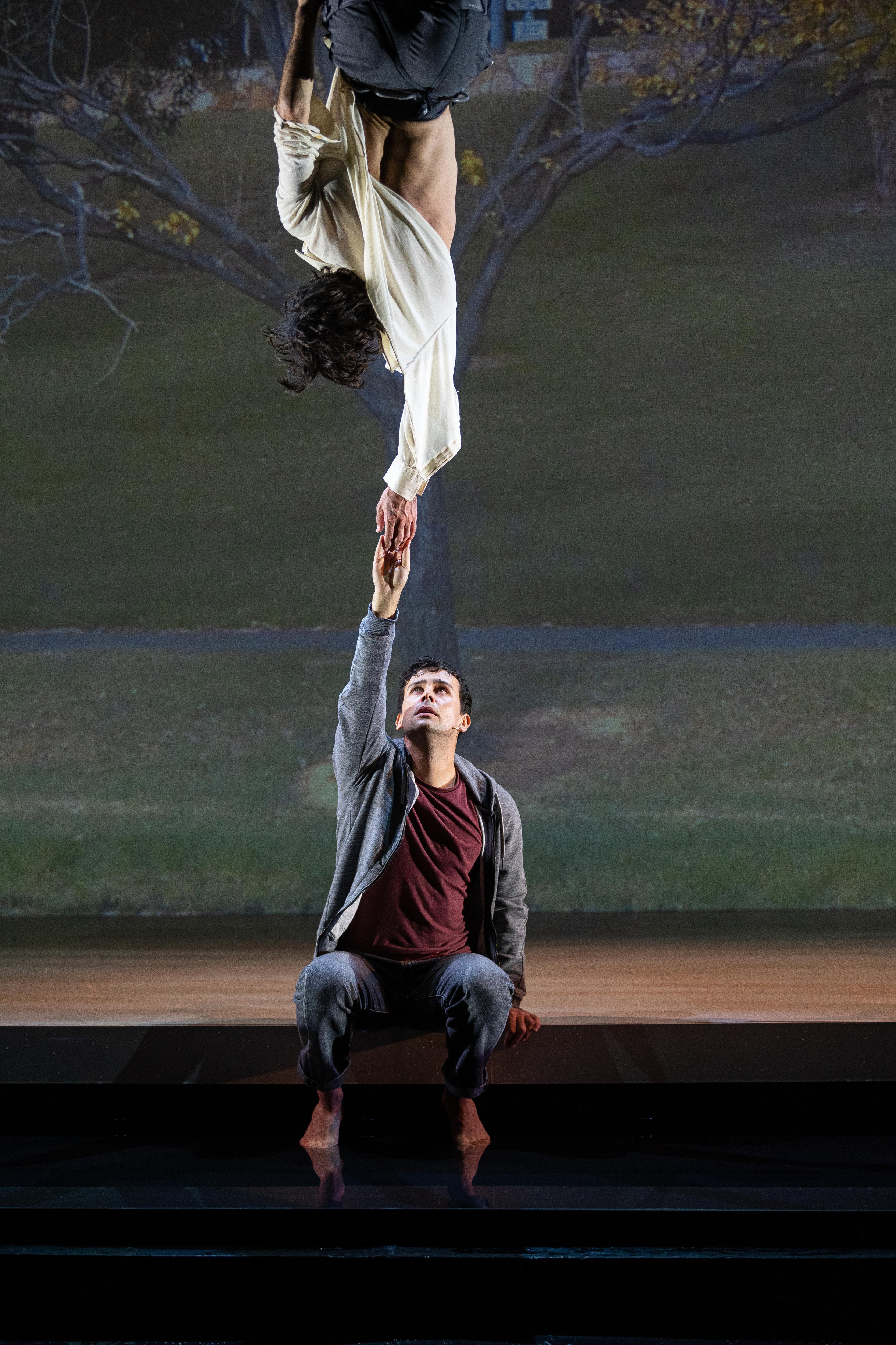 A young white man sits onstage and reaches up to touch the hand of a young white male dancer, suspended from the ceiling.