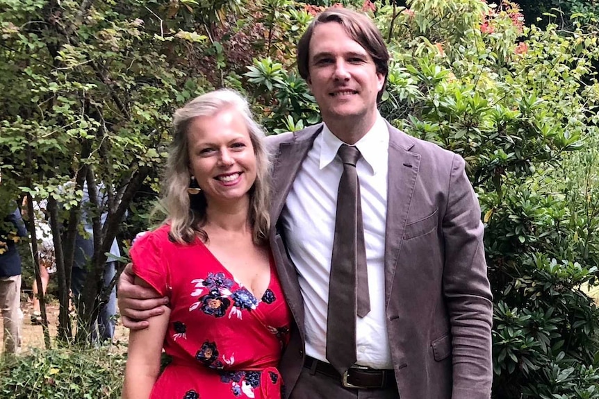 A woman and a man stand facing the camera with their arms around one another, smiling.