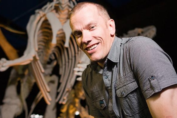 A man in a grey shirt smiles front of whale skeleton at a museum