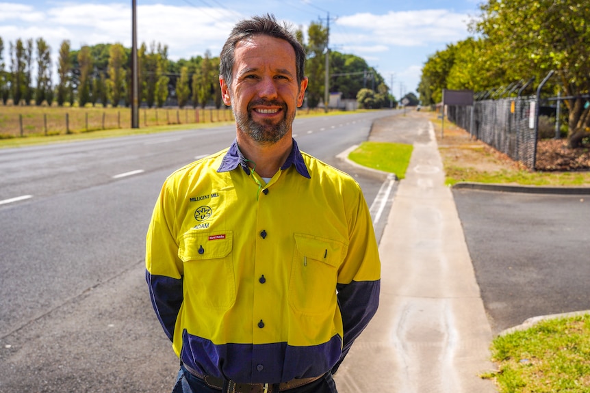 A man in a high-vis shirt smiles at the camera with a road behind him