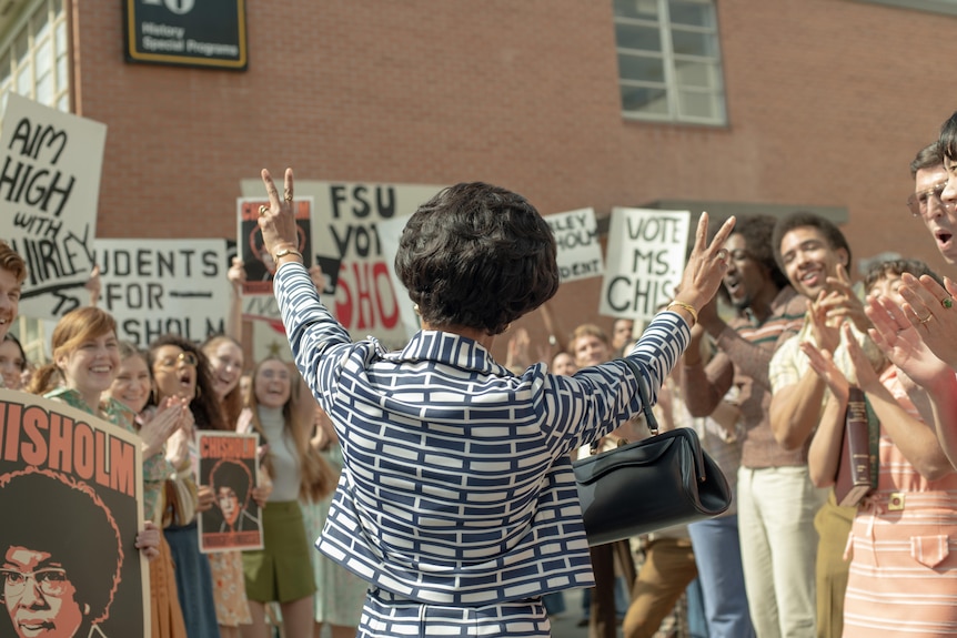 Regina has her back to the camera and stands holding both hands up with peace signs in front of happy protesters.