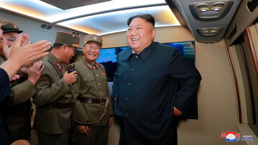 Kim Jong-un in front of a screen laughing while military leaders clap and laugh.