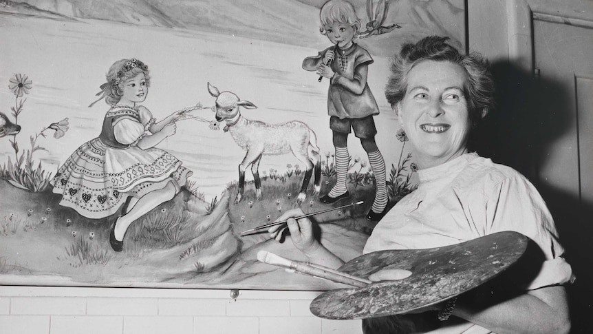 A black and white photo of artist Pixie O'Harris from about 1951. She's holding a palette, brushes and painting while smiling.