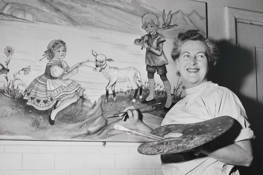 A black and white photo of artist Pixie O'Harris from about 1951. She's holding a palette, brushes and painting while smiling.