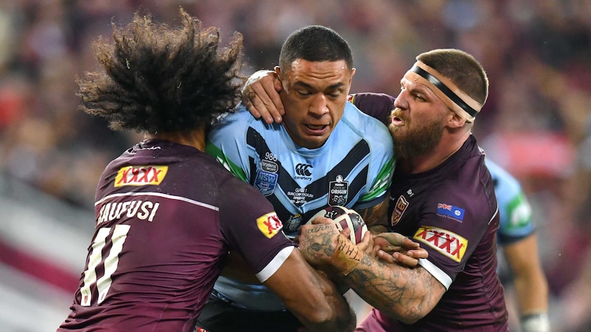 A New South Wales State of Origin player holds the ball as he is tackled by two Queensland players on his left and right.