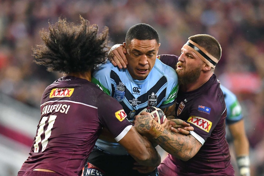 A New South Wales State of Origin player holds the ball as he is tackled by two Queensland players on his left and right.