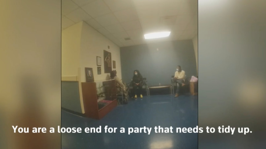A still image from a police officer's bodycam shows three people meeting at the end of a room under a light.