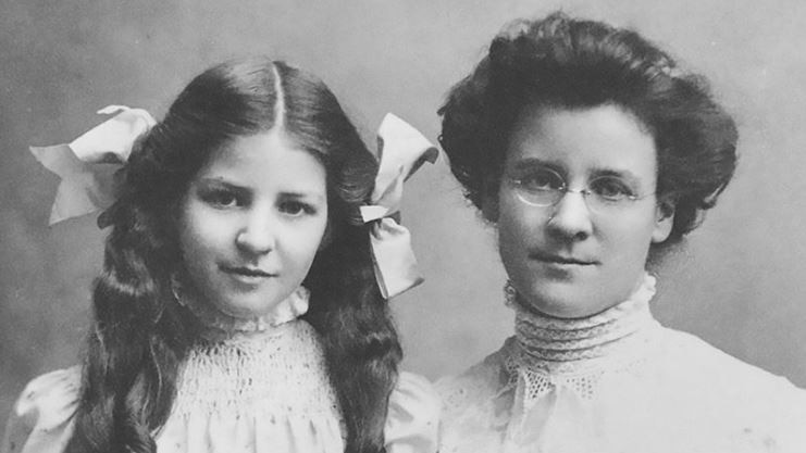 Black and white image of girl with bows in her long hair, sitting with woman wearing glasses, both with neutral expressions.