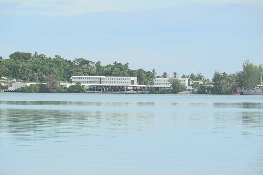 Beyond a body of water sit a number of buildings surrounded by trees.