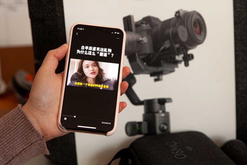 You view a woman's hand holding an iPhone beside camera equipment showing a woman of Asian descent pictured on the screen.