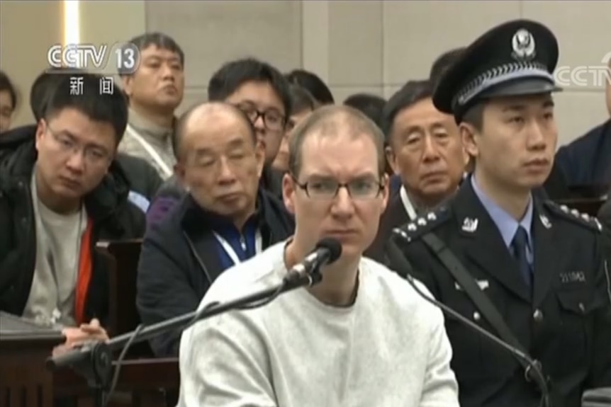 A man wearing a grey sweatshirt sits in front of two microphones.