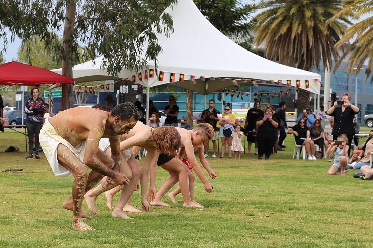 A line of young men dance along a grassed area as an audience watches on.