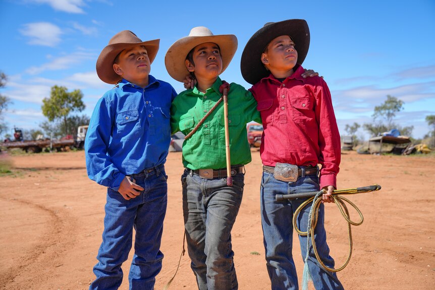 Three young boys dressed as stockmen stand arm in arm, looking up to the sky.