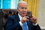 An elderly white man in a suit and blue tie gestures with his thumbs and index fingers