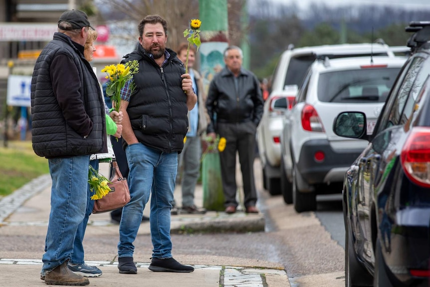 Man standing on footpath holding sunflower, other people around him holding yellow flowers.