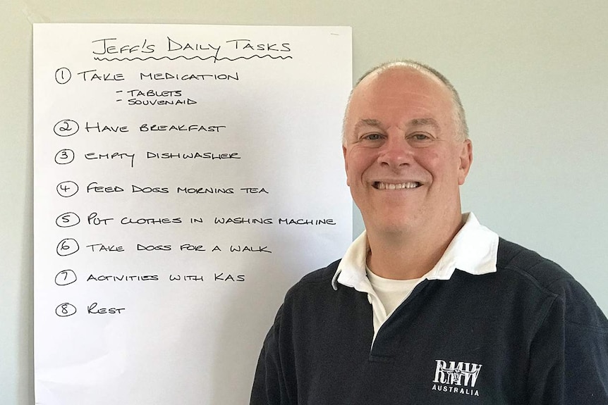 Jeff Thurlow, who has younger onset dementia, smiles as he stands beside a list on the wall to remind him of his daily tasks.
