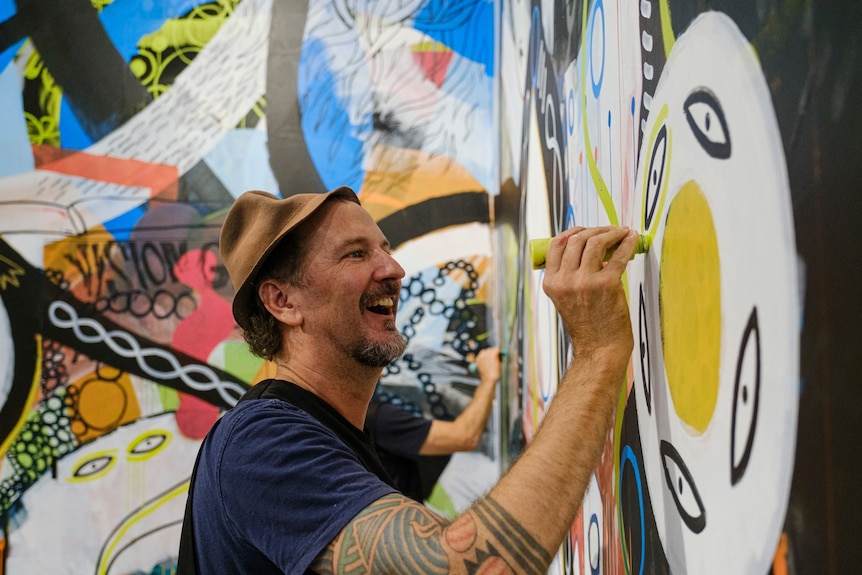 Man wearing a hat paints a colourful picture while laughing.