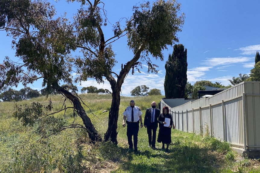 Two men in suits and a woman holding papers as they walk through an empty block of land beside a fence