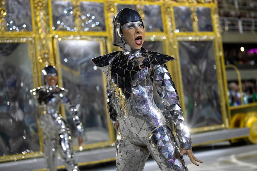 a dancer in a silver suit and headwear with silver make up over her eyes dances beside a float at Carnival in Rio