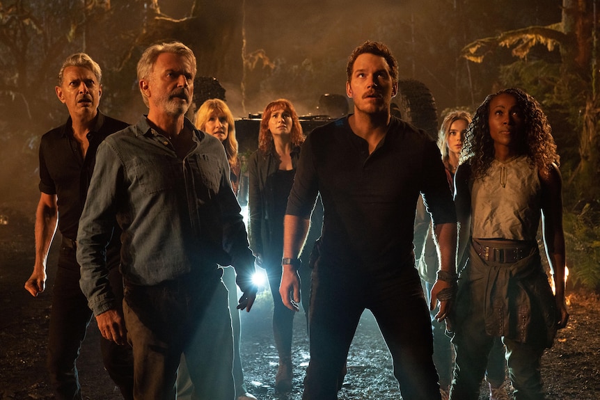 Seven people stand braced with anticipation and looking upwards at a dinosaur that is off-camera in a jungle setting at night.