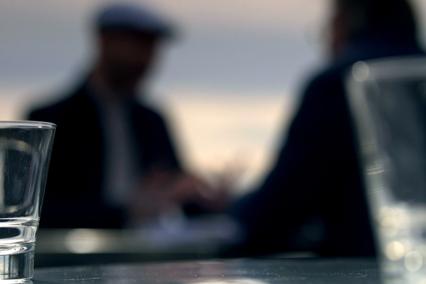 Two people sit at a table. They are out of focus. In the foreground there are two glasses on a table.
