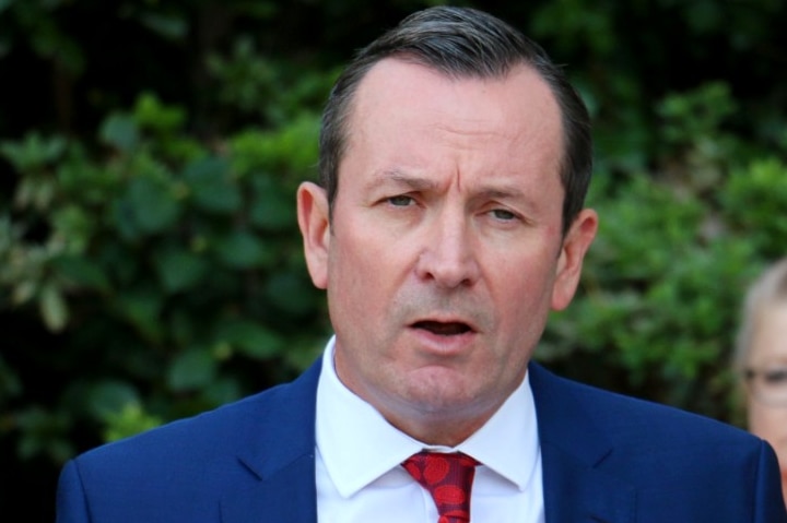 Mark McGowan wearing a red tie and blue suit stands in front of a bank of microphones with Sue Ellery standing behind him.