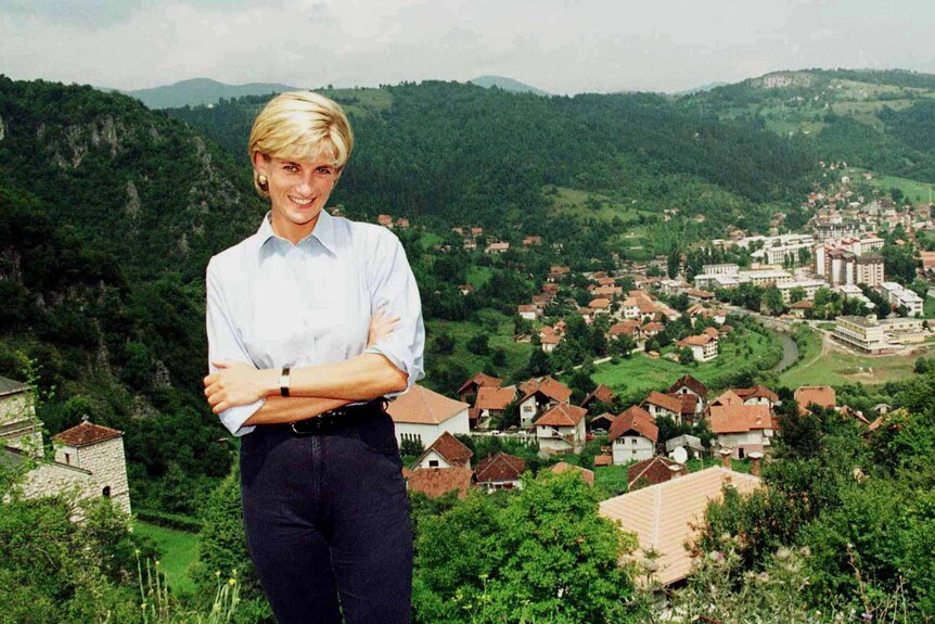 Princess Diana smiles while standing on top of a plateau with a Bosnian village nestled between green hills in a valley below.