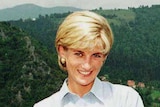 Princess Diana smiles while standing on top of a plateau with a Bosnian village nestled between green hills in a valley below.