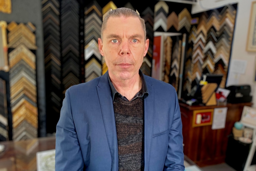 Mark Heydon standing in his shop in Burwood, Melbourne with a selection of frames behind him.