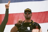 A man dressed in black and yellow tactical gear smokes a cigarette in front of an American flag