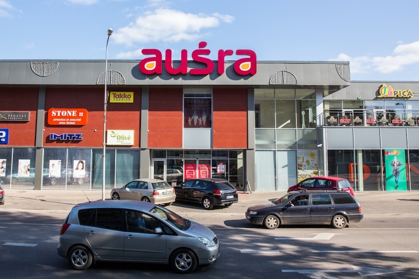 The facade of a shopping centre in Lithuania, with cars parked in the foreground.