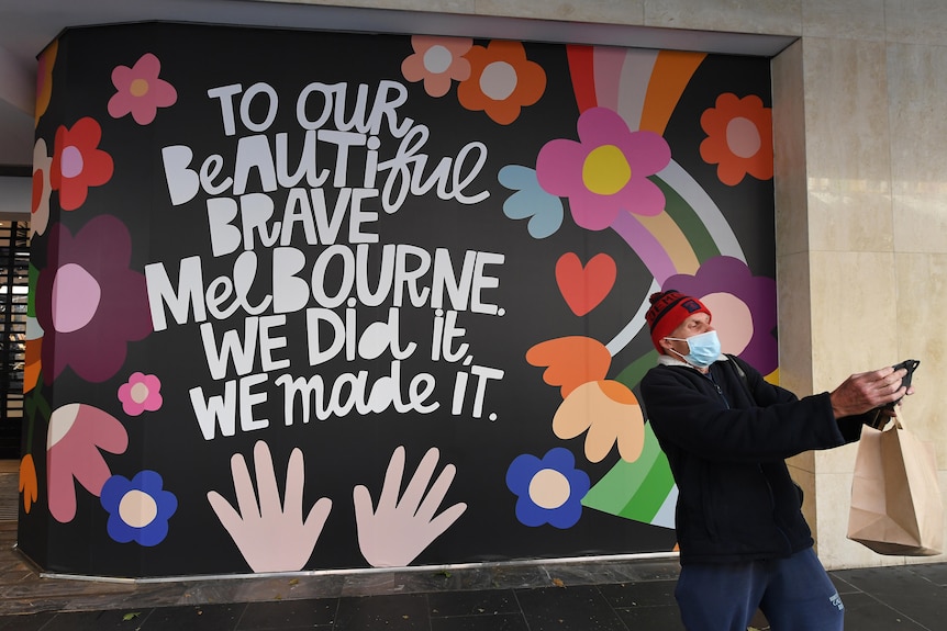 A man takes a selfie in front of a colourful wall with the words 'to our beautiful brave melbourne we did it we made it' on it