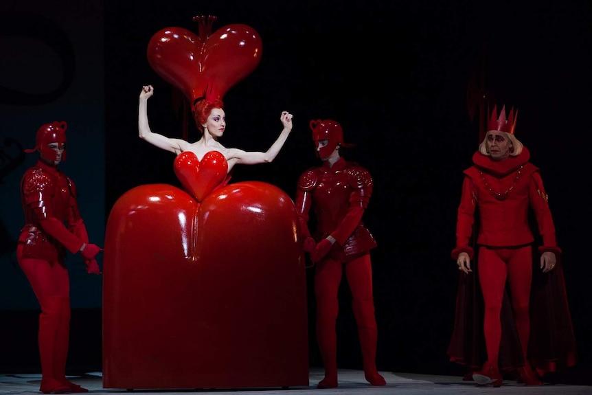 A ballet dancer on stage in a giant red heart costume, backed by three other performers dressed in red.