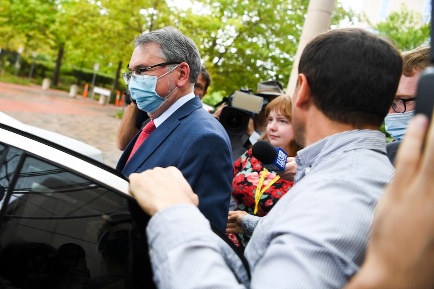 A man in a suit with grey hair, wearing a mask, enters a car.